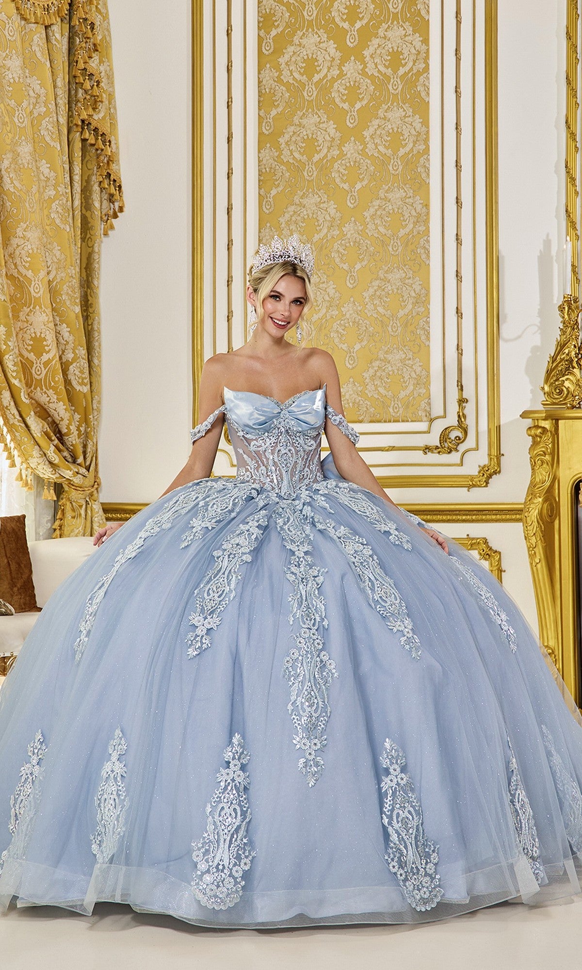 A woman in the Quinceanera Dress 5087 by Dancing Queen, a dusty blue off-the-shoulder gown with ornate details and a tiara, stands in an opulent, gold-accented room with patterned wallpaper and luxurious curtains.